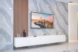 A modern living room with a TV mounted on a marble wall, a white console table with decorative items, and a view of a coastal scene. - Spacious Family Retreat w/ Pool at Windsor Island (Davenport)