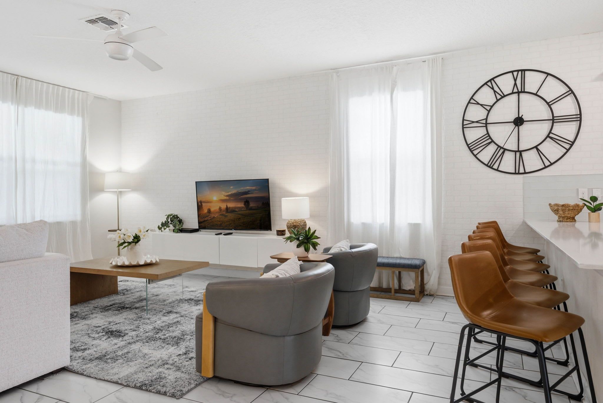 Una sala de estar moderna con sofá, sillones, una mesa de centro, televisor, un reloj de pared grande y una zona de bar con taburetes.