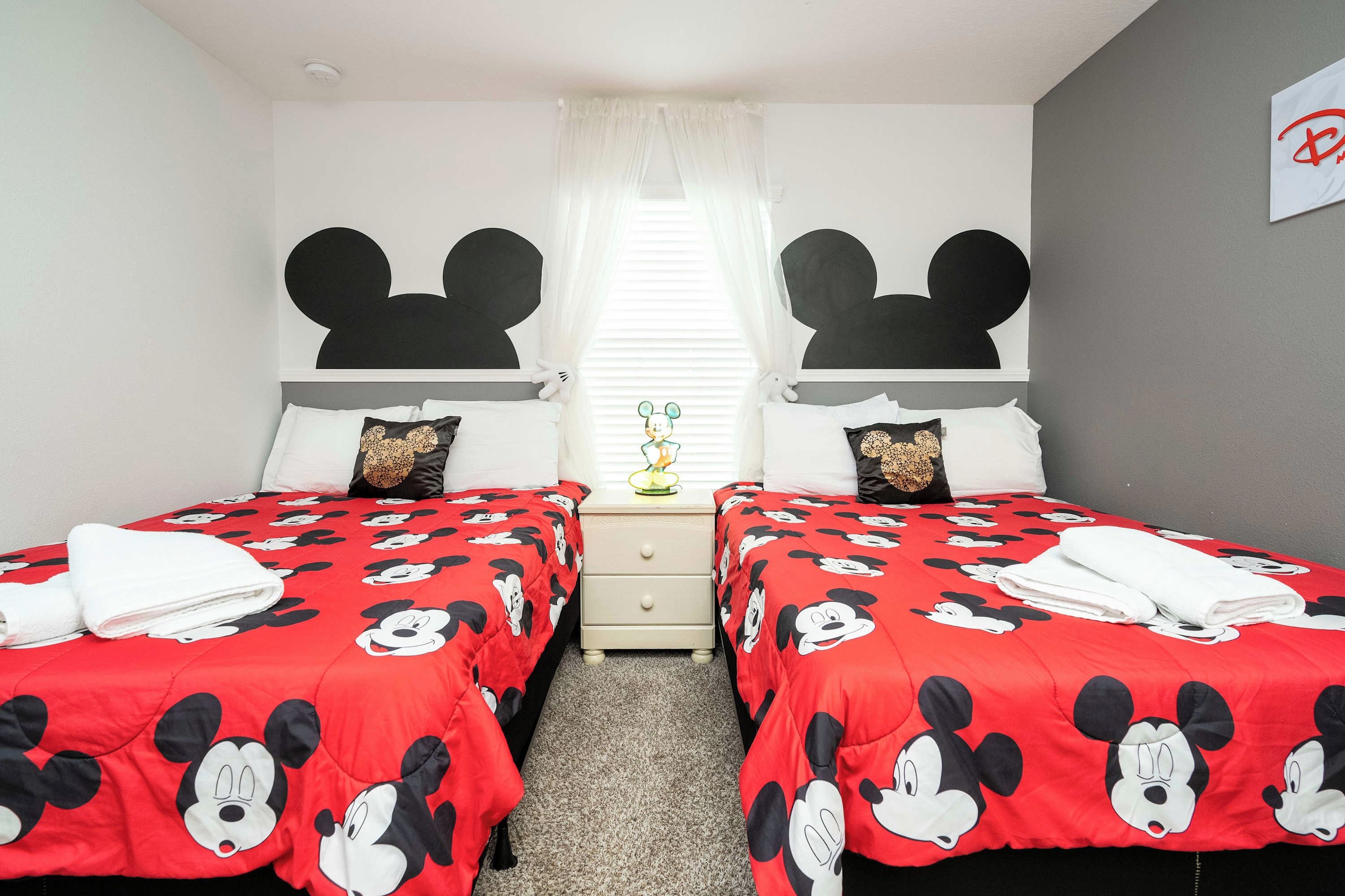 A hotel room with two beds, each with a Mickey Mouse themed bedspread. There is a nightstand between the beds with a lamp, and a Mickey Mouse decoration on the wall.