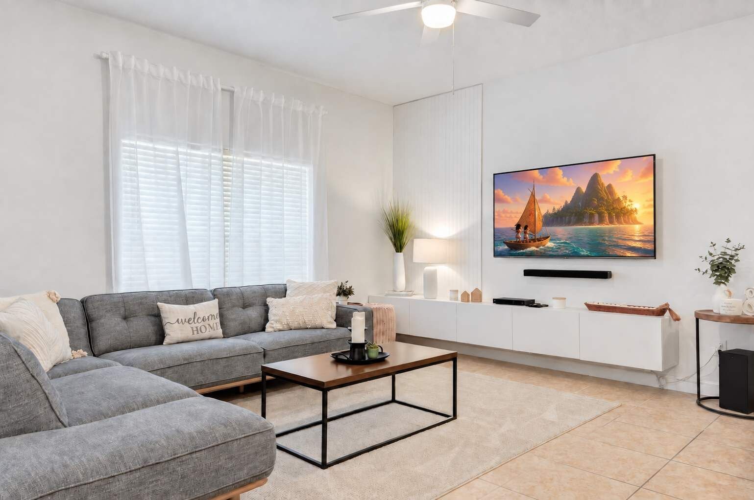 A modern living room with a grey sectional sofa, a coffee table, a TV mounted on the wall, and a window with white curtains.