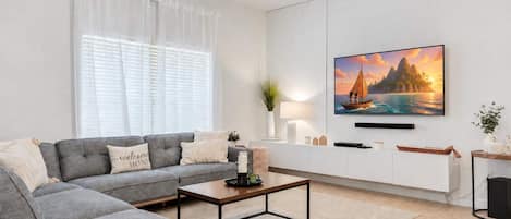A modern living room with a grey sectional sofa, a coffee table, a TV mounted on the wall, and a window with white curtains.
