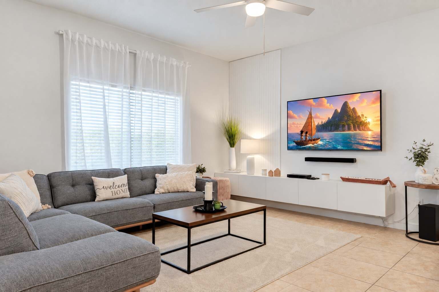 A modern living room with a grey sectional sofa, a coffee table, a TV mounted on the wall, and a window with white curtains.