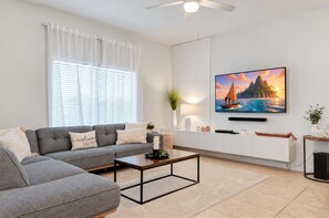 A modern living room with a grey sectional sofa, a coffee table, a TV mounted on the wall, and a window with white curtains. - Family-friendly Townhouse With Pool, Minutes to Disney! (Kissimmee)