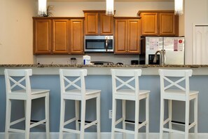 A kitchen with wooden cabinets, a stainless steel microwave, a refrigerator, and three white bar stools. - Family-friendly Townhouse With Pool, Minutes to Disney! (Kissimmee)