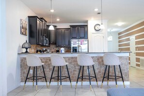 A modern kitchen with dark cabinets, a central island with bar stools, a clock, and a pendant light. - Spacious Family Retreat w/ Game Room & Steps to Clubhouse (Davenport)