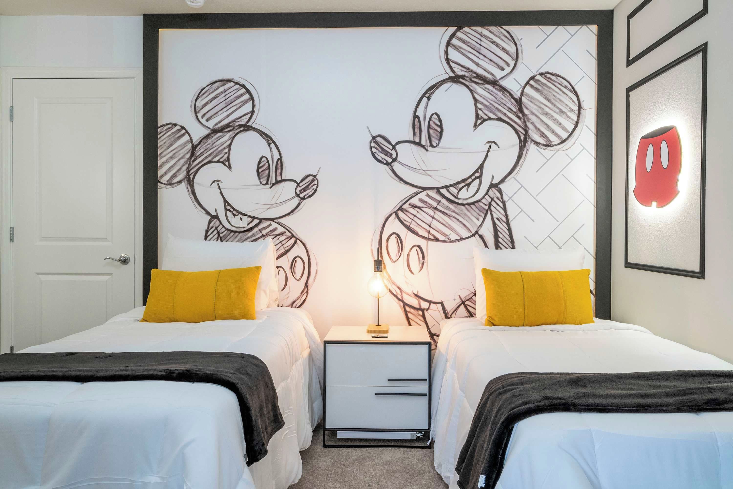 Habitación de hotel con dos camas, una mesita de noche, una pared con dibujos de Mickey Mouse y una luz roja en la pared.