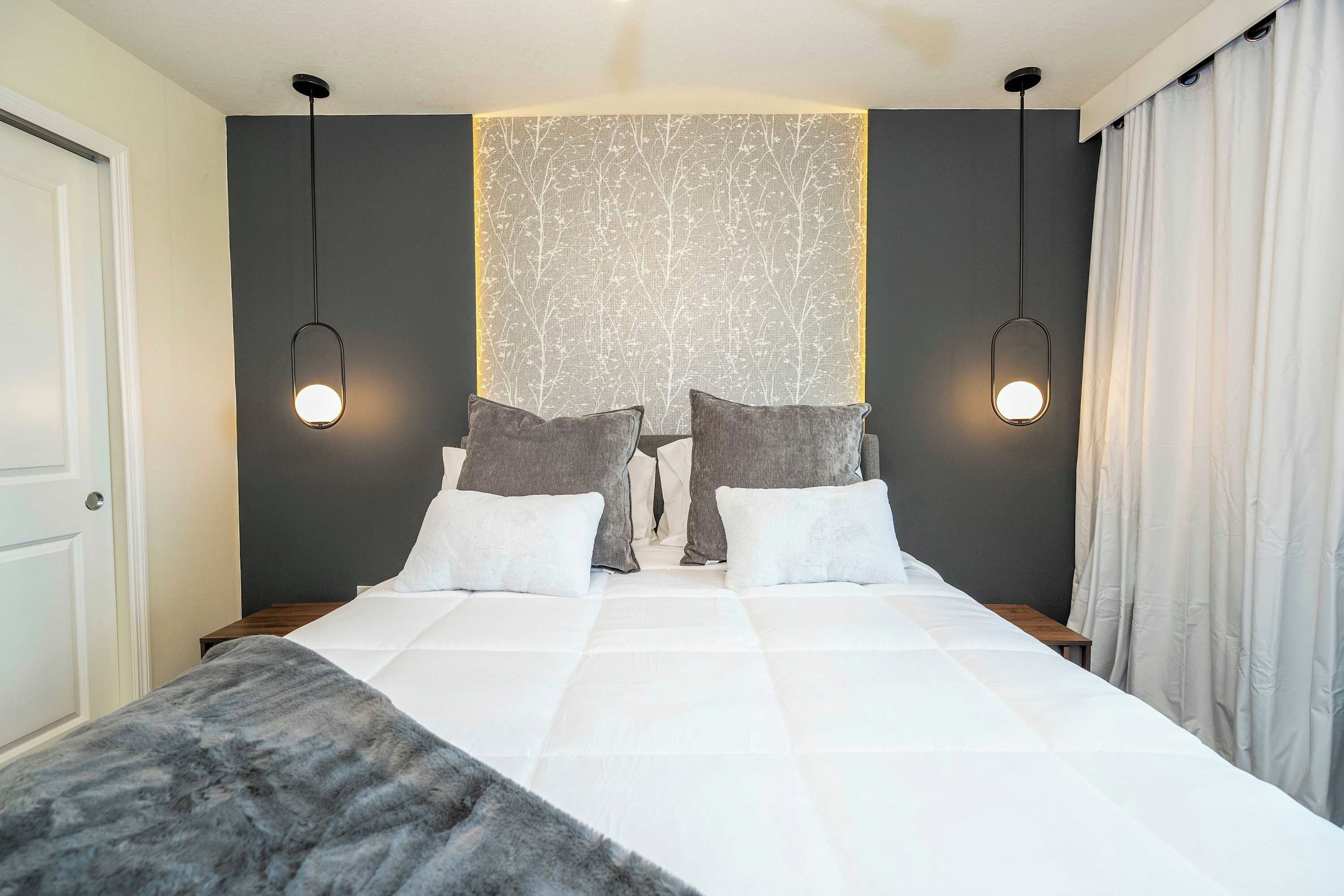 Una cama bien hecha con ropa de cama blanca, dos almohadas grises y una manta gris. Hay dos mesitas de noche con lámparas, un ventanal con cortinas y una pared con un diseño texturizado.