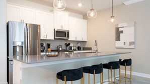 Een moderne keuken met witte kastjes, roestvrijstalen apparatuur, een centraal eiland met barkrukken en hanglampen.