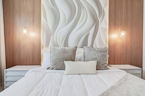 Ein Schlafzimmer mit modernem Design, mit einem großen Bett mit weißer Bettwäsche und mehreren Kissen, einer dekorativen Wandverkleidung und zwei Nachttischlampen.