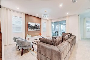 Ein modernes Wohnzimmer mit einem großen Ecksofa, einem kleinen Sessel, einem flachbildfernseher an der Wand und einer Glastür zum Balkon.