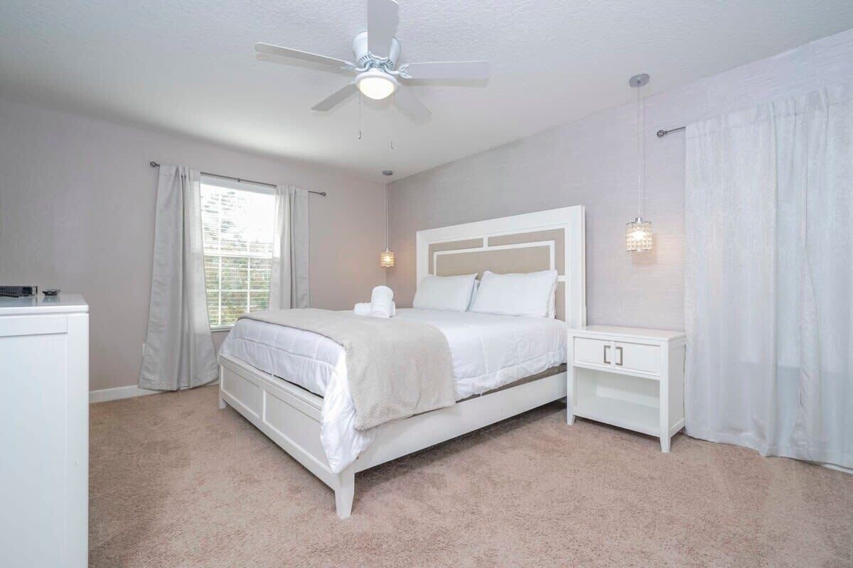 Une chambre à coucher avec un lit, des tables de chevet, un ventilateur de plafond et une fenêtre avec des rideaux.