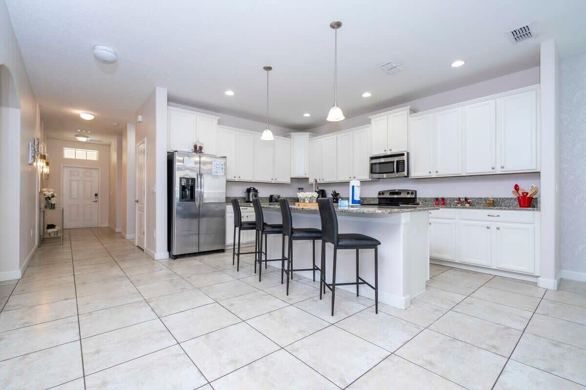 A modern kitchen with white cabinets, a stainless steel refrigerator, a microwave, and a granite countertop. There are three black bar stools and pendant lights above the island.