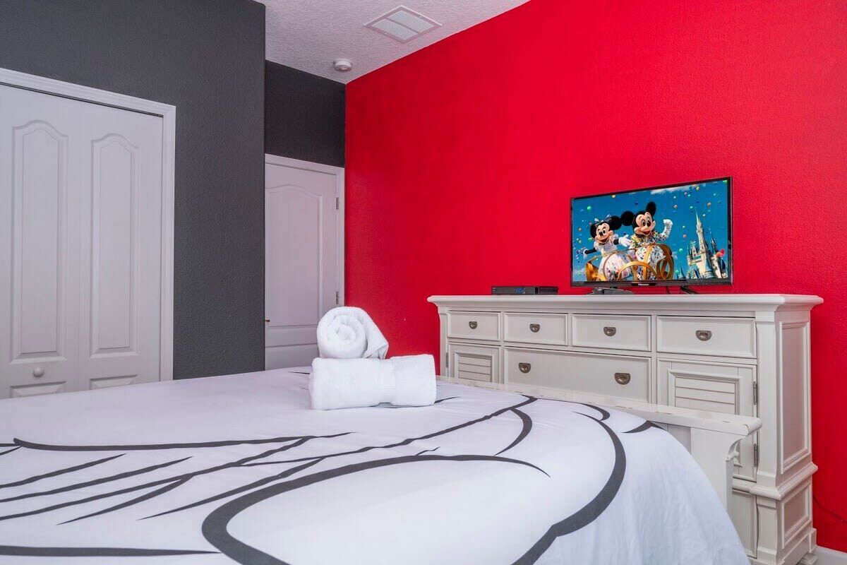 Een hotelkamer met een bed, een witte dressoir, een rode accentmuur en een schilderij van Mickey Mouse.