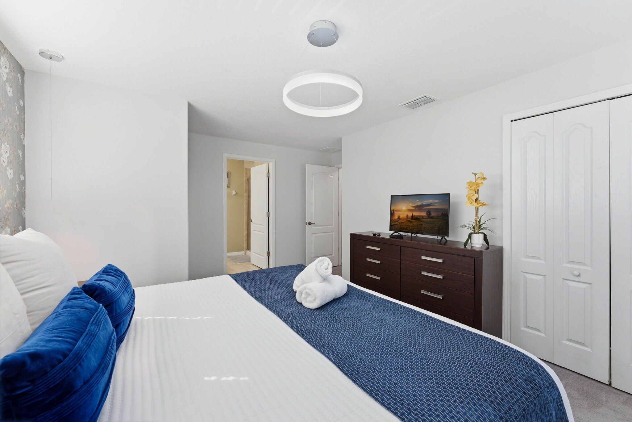 Ein modernes Hotelzimmer mit einem großen Bett, einer dunklen Holzkommode, einem weißen Kleiderschrank und einem Flachbildfernseher auf einem Standfuß.