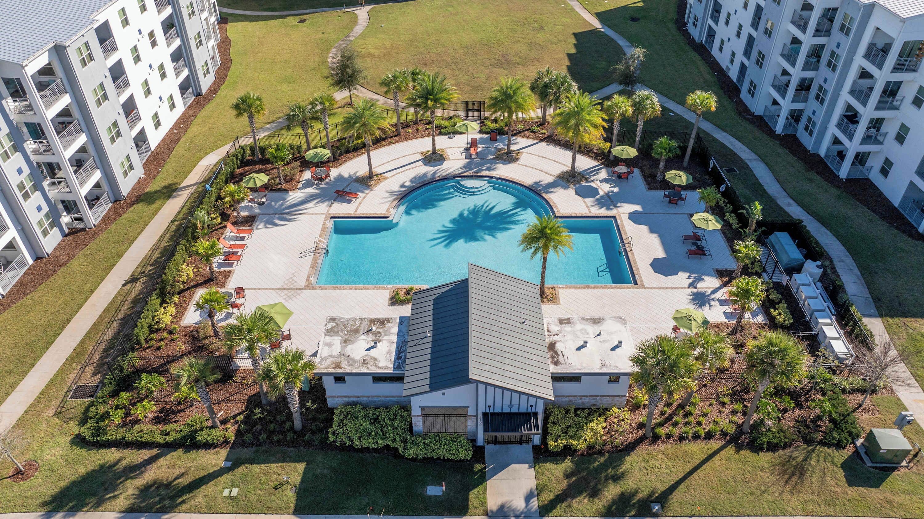 An aerial view of a hotel with a central swimming pool, surrounded by palm trees and lounge areas.