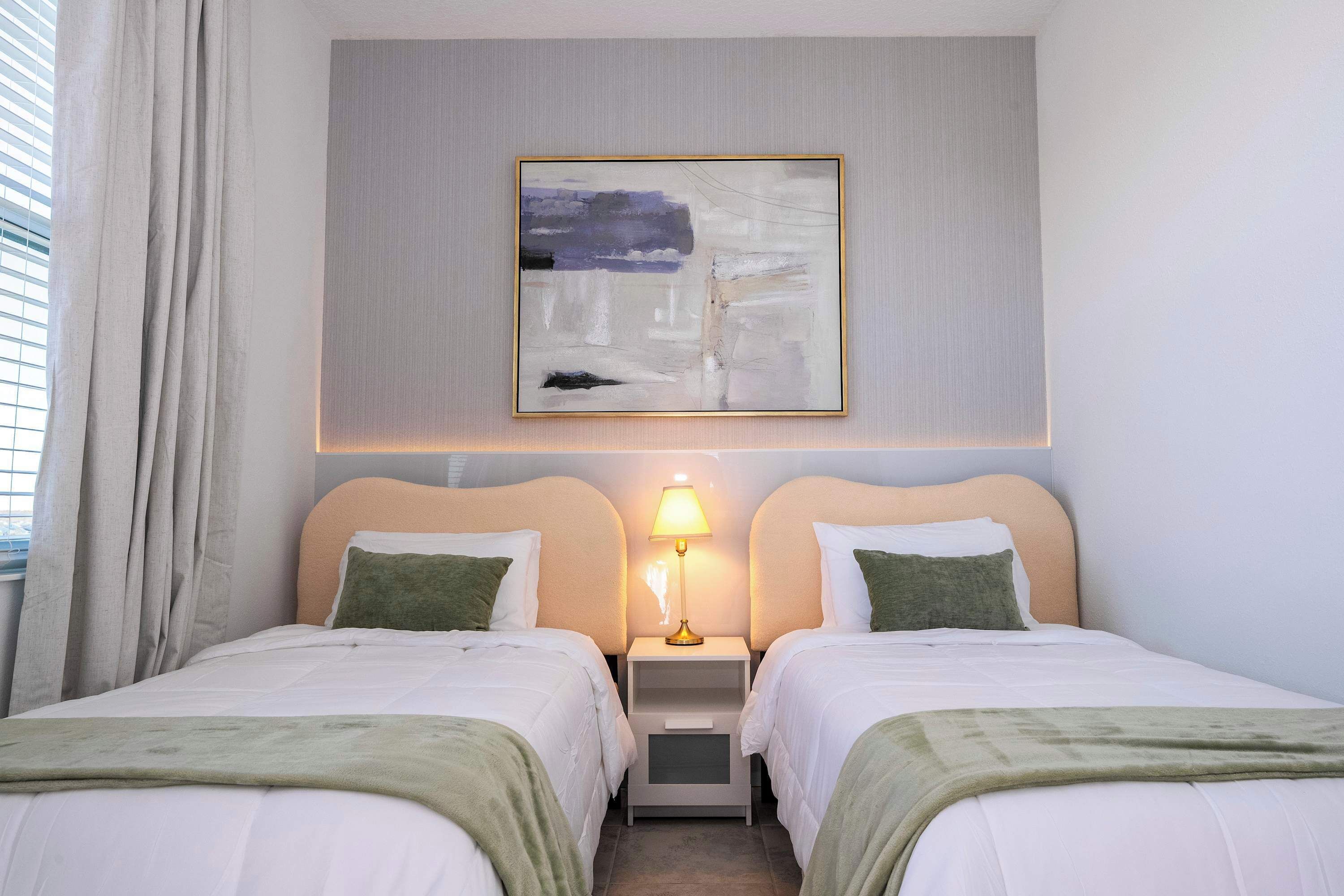 A hotel room with two beds, a nightstand with a lamp, a painting on the wall, and a window with curtains.