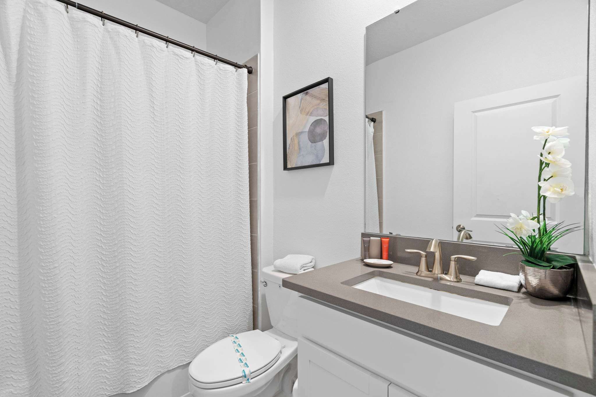 A modern bathroom with a white toilet, a beige sink with gold faucets, a white shower curtain, and a potted orchid.