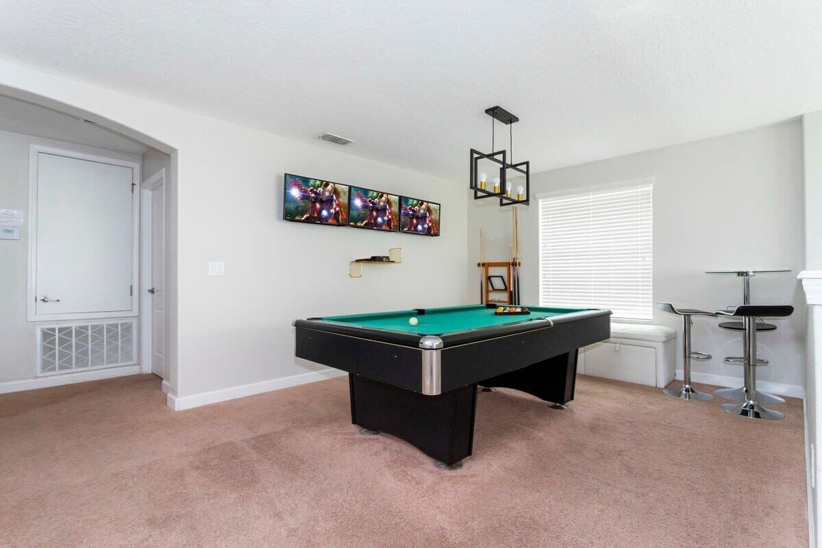 A pool table in a room with a white door, a window with blinds, and a bar area with stools.