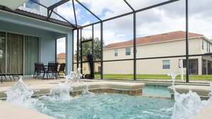 A swimming pool with water jets, a covered patio with chairs, and a view of houses in the background. - Charming Resort House w/ Private Pool & Fast Wifi (Kissimmee)