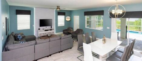 A modern living room with a sectional sofa, a flat-screen TV, and a dining area with a white table and chairs.