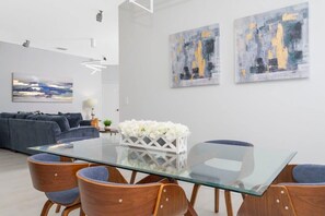 Ein modernes Wohnzimmer mit einem Esstisch aus Glas, Holzstühlen, einem grauen Sofa und abstrakten Bildern an der Wand.