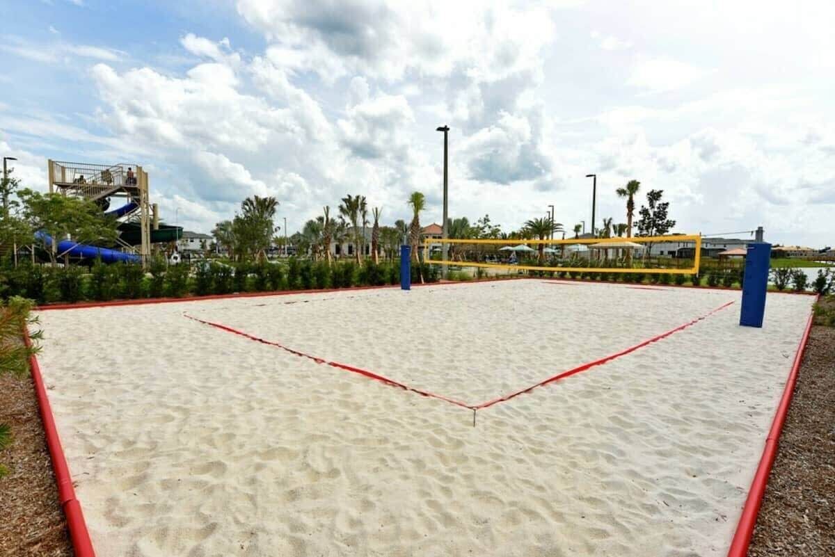 Una cancha de voleibol de arena con una red, rodeada de palmeras y un tobogán de agua al fondo.