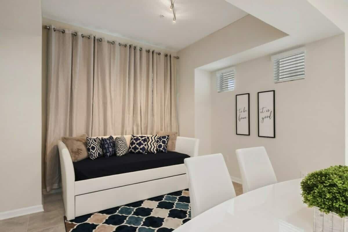 Ein modernes Wohnzimmer mit Sofa, Esstisch und Stühlen. Vorhänge, gerahmte Bilder an der Wand und eine kleine Pflanze auf dem Tisch sind ebenfalls zu sehen.