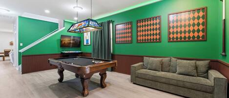 Een biljart tafel in een ruimte met groene muren, een bank en kunstwerken in lijsten.