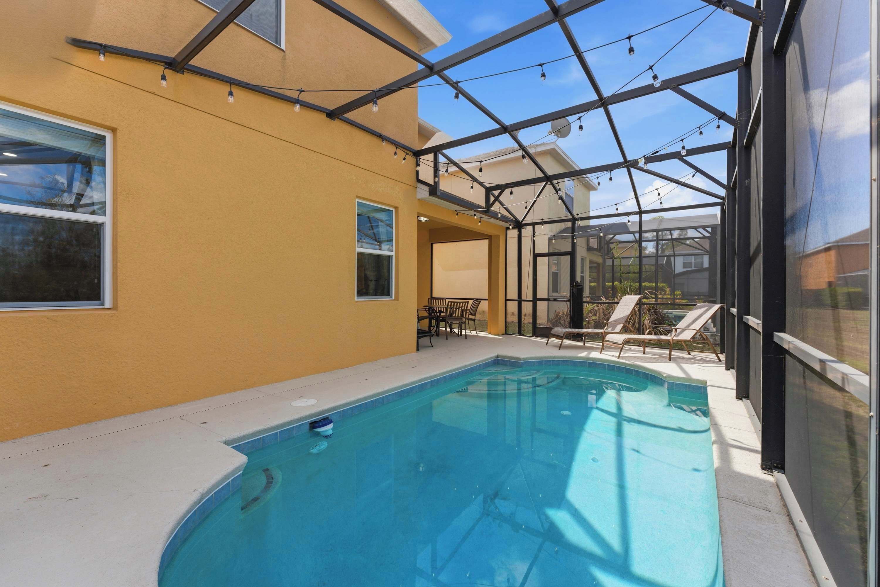 A swimming pool with a glass enclosure, a patio area with chairs, and a clear sky.