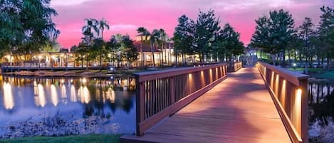 Un puente de madera cruza un cuerpo de agua tranquilo, rodeado de árboles y edificios con iluminación cálida.