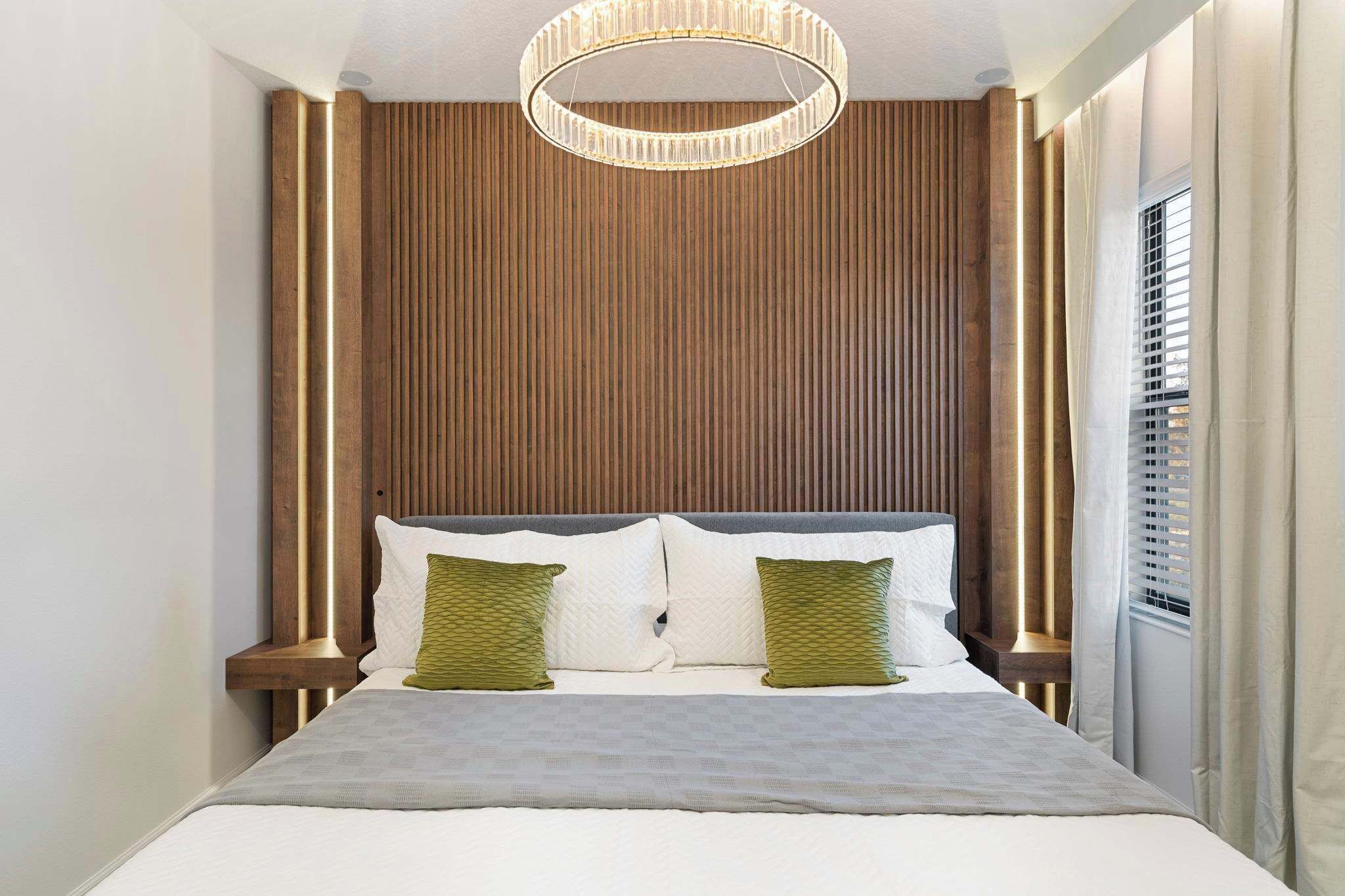 Una camera da letto moderna con una testiera in legno, un letto grande con biancheria da letto bianca, due cuscini verdi e una lampada a sospensione circolare.