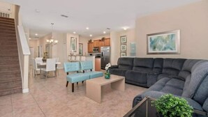 A living room with a gray sofa, a coffee table, and a blue armchair. There is a staircase on the left and a kitchen in the background. - Family-friendly Paradise Palms Townhouse With Pool Access (Kissimmee)