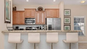 A kitchen with wooden cabinets, a microwave, a refrigerator, and a bar area with four stools. - Family-friendly Paradise Palms Townhouse With Pool Access (Kissimmee)