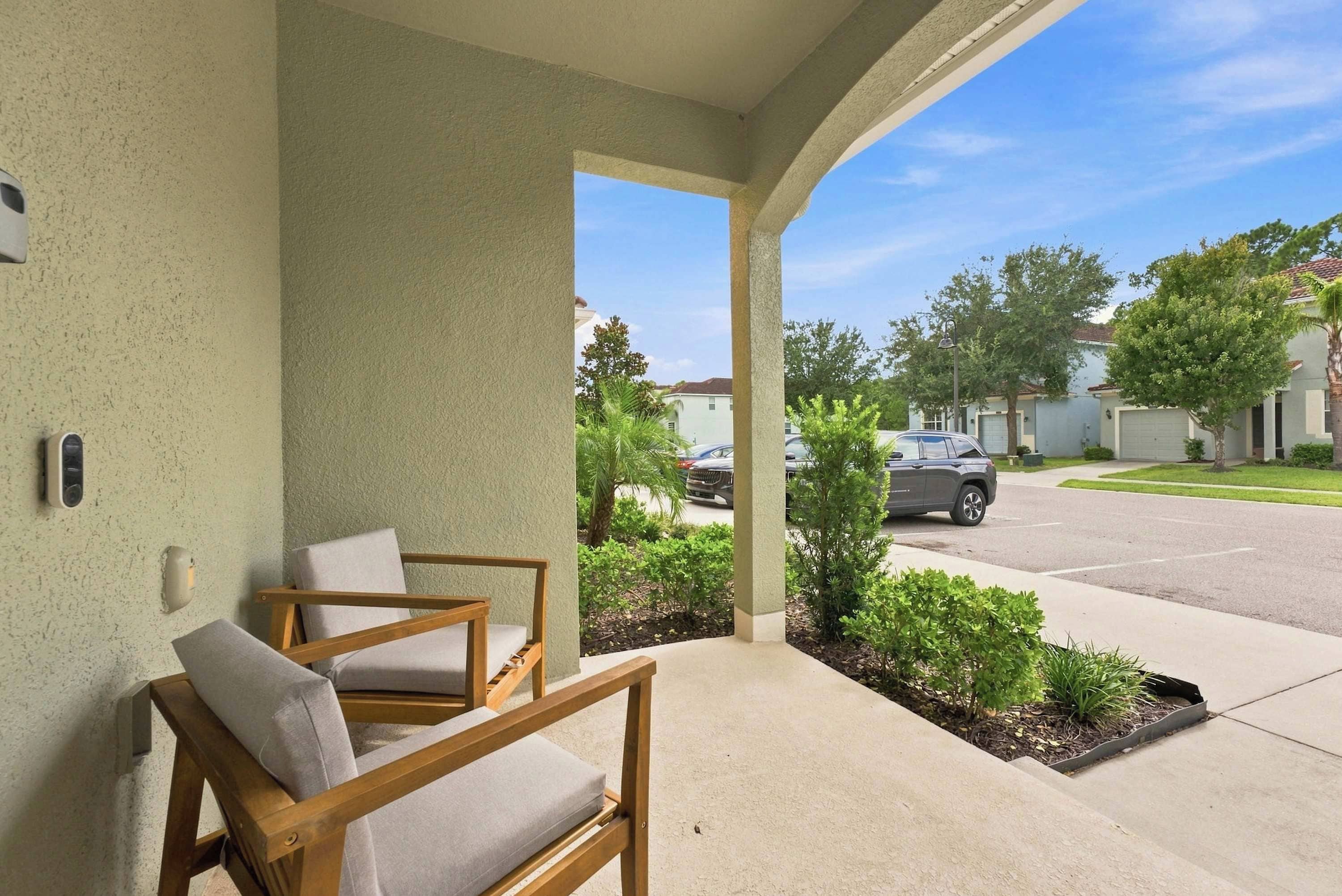 A covered porch with two wooden chairs and a small table, a clear view of a residential street, and a well-maintained lawn.