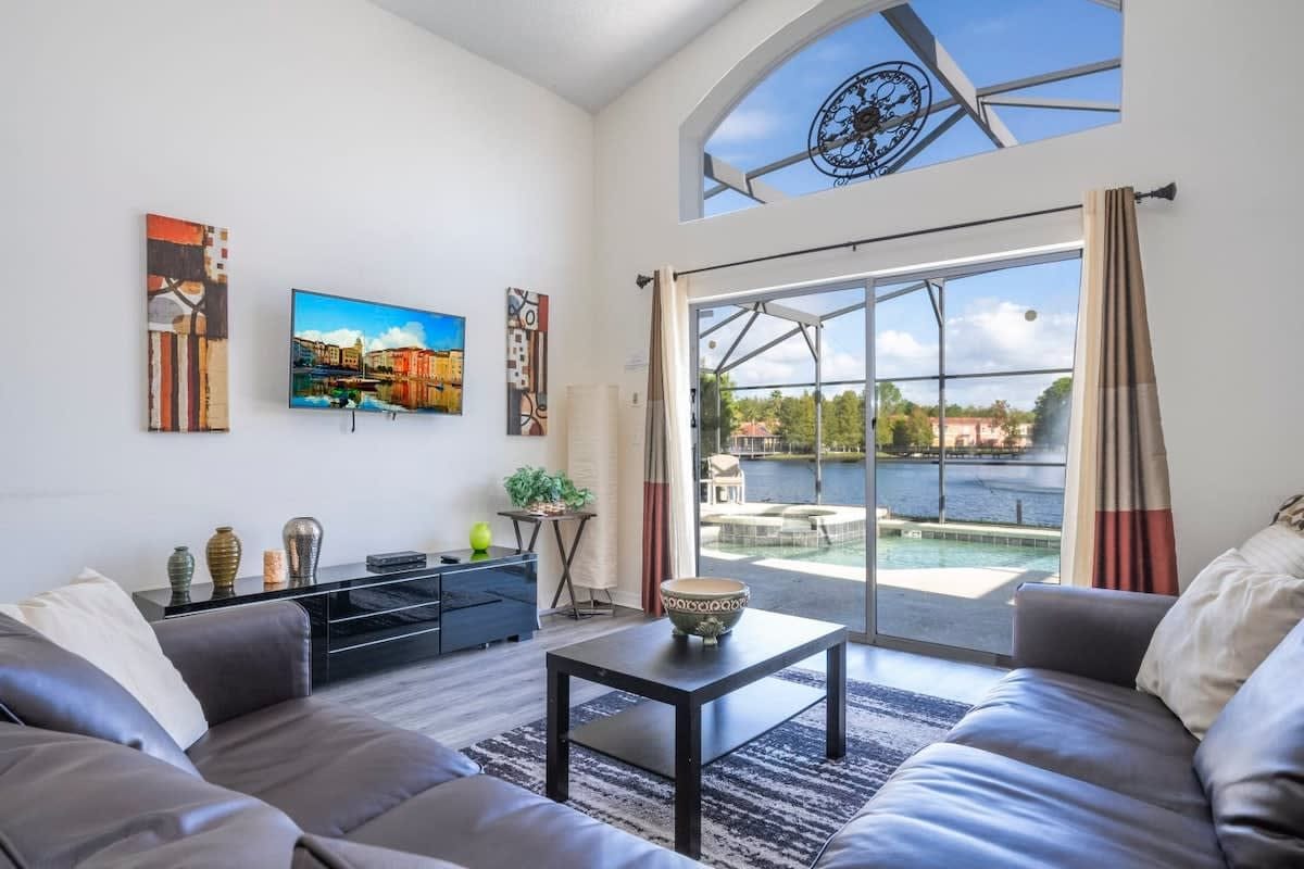 A modern living room with a sofa, coffee table, TV, artwork, and a large window overlooking a body of water.