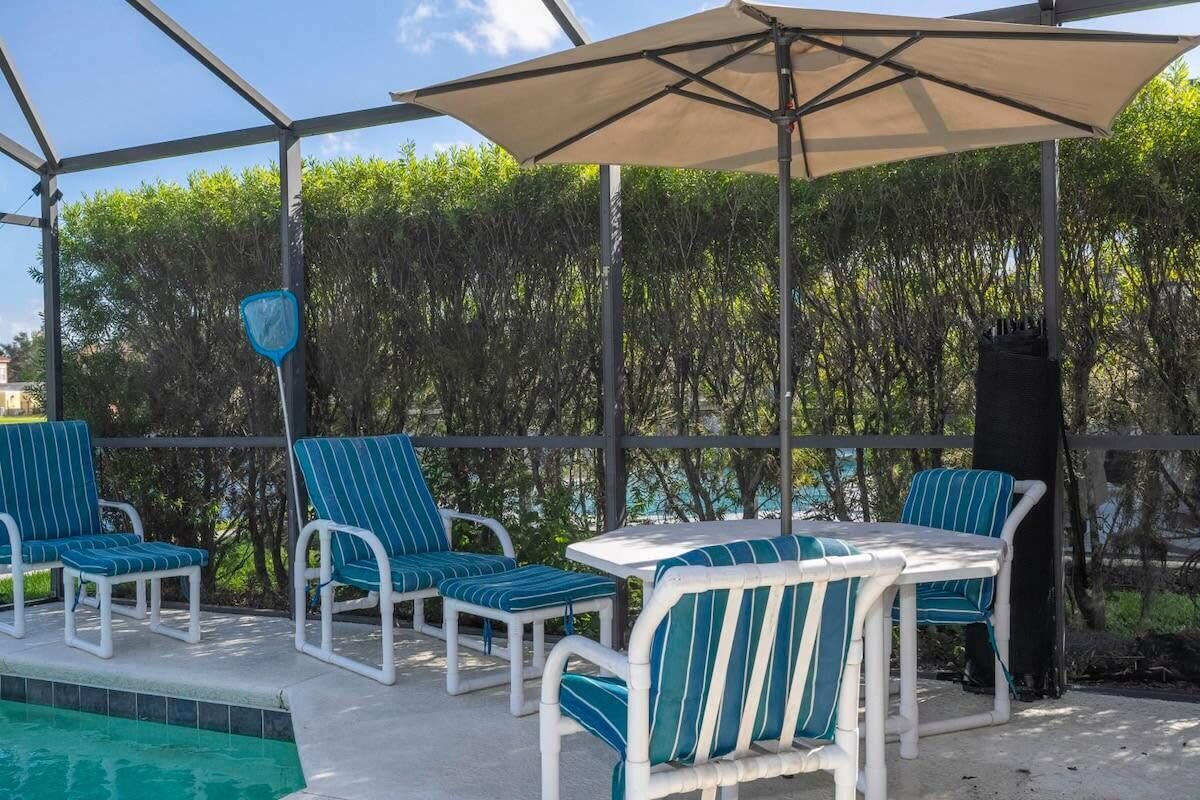 Uma área externa coberta com espreguiçadeiras e uma mesa, cercada por uma piscina e vegetação.