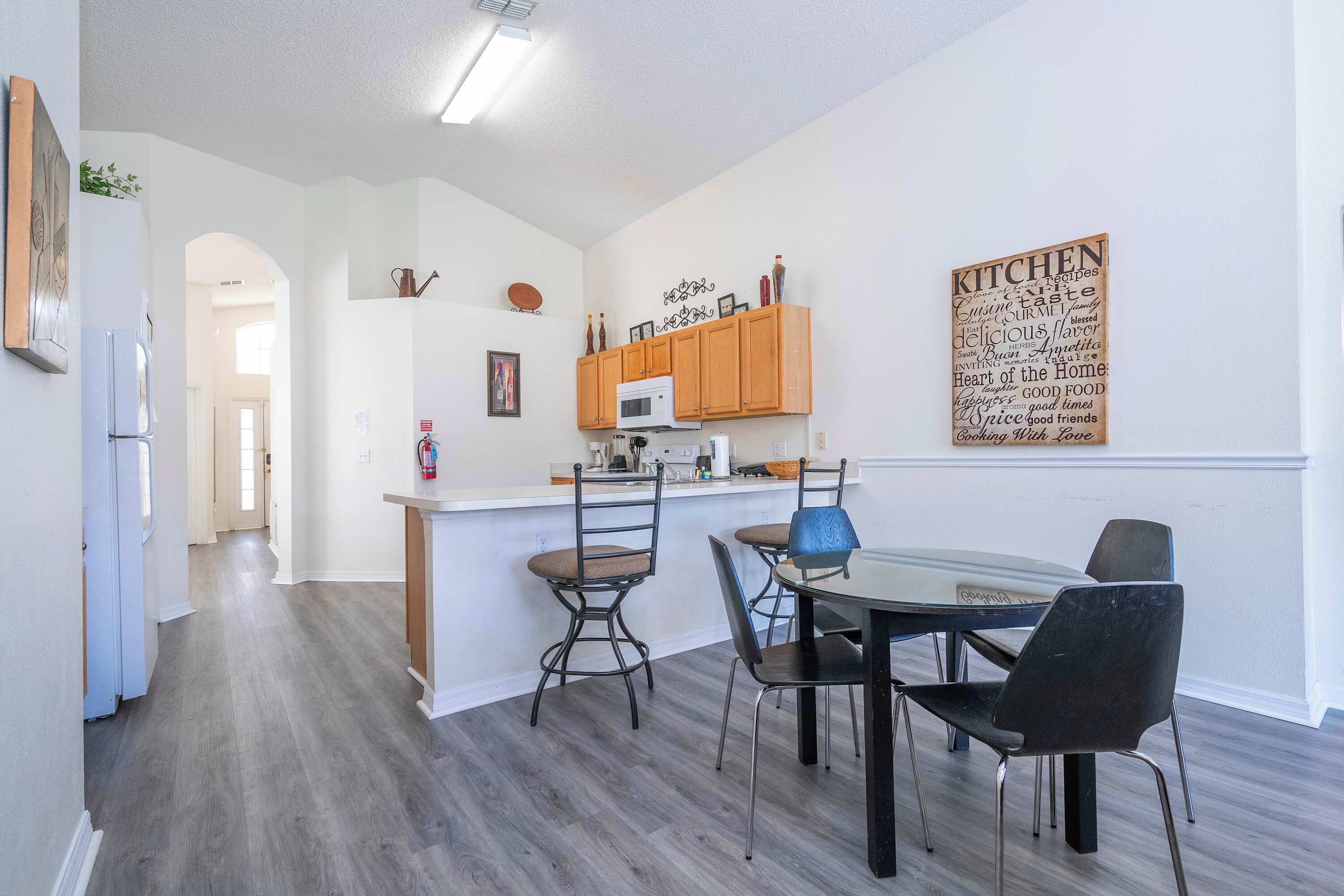 Una cocina moderna con armarios de madera, un microondas y una nube de palabras en la pared. Hay un comedor con una mesa de vidrio y sillas.
