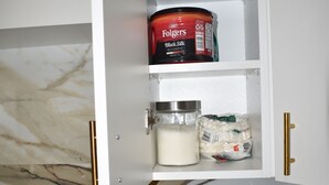 A kitchen with a marble backsplash, a coffee maker, and a cabinet containing Tim Hortons coffee, coffee whitener, and black silk coffee creamer.