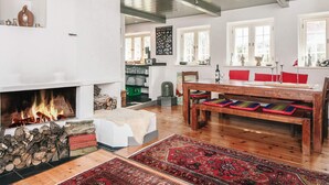 Een ruime woonkamer met een open haard, houten vloeren, een eethoek met rode kussens en een groot raam met veel ruitjes.