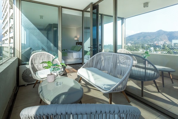 Ein Balkon mit Korbmöbeln, einem Glastisch und Blick auf eine Stadtlandschaft und Berge.