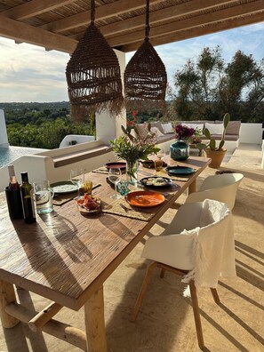 Uma área de refeições ao ar livre com mesa de madeira, luminárias de vime pendentes, vista para vegetação e uma planta de cacto.