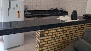 A kitchen with a black countertop, a white refrigerator, a brick accent wall, and a grey couch. - Pousada Apart Praia Uruau (Beberibe)
