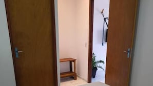 A bedroom with a bed, two wooden doors, a small wooden bench, and a potted plant in the background. - Pousada Apart Praia Uruau (Beberibe)
