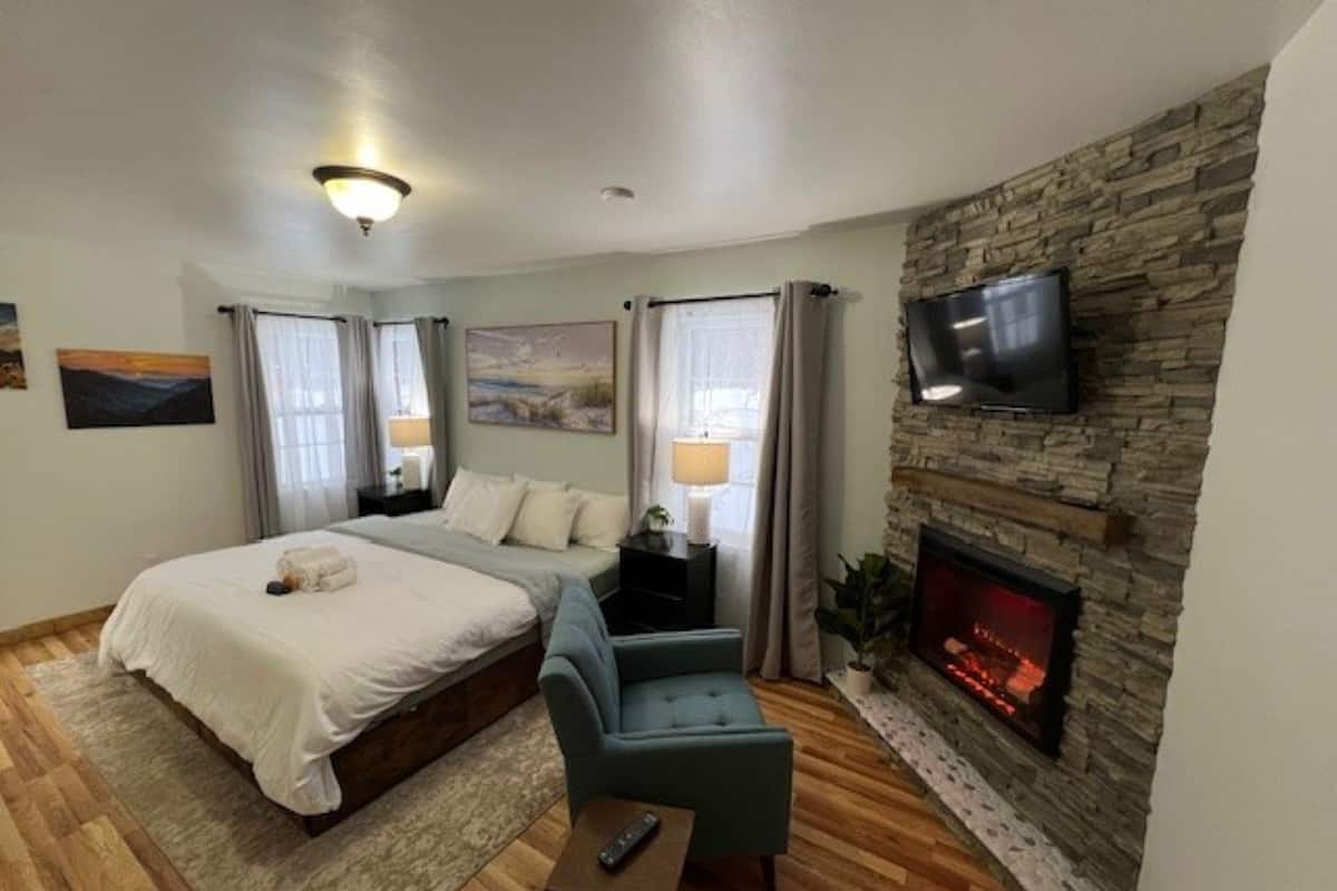 A bedroom with a bed, a TV mounted on a stone wall, a fireplace, and a small seating area.