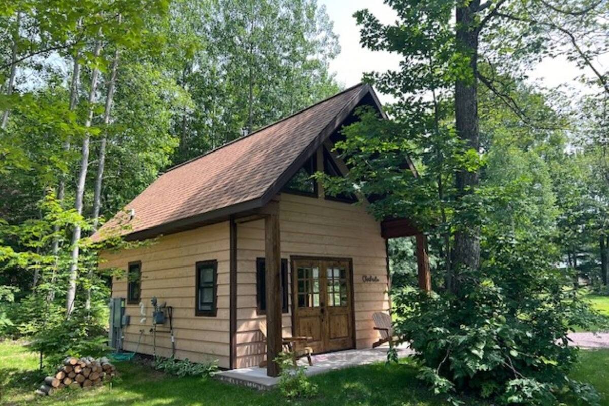 A small, single-story wooden cabin with a sloped roof, surrounded by lush greenery and trees.