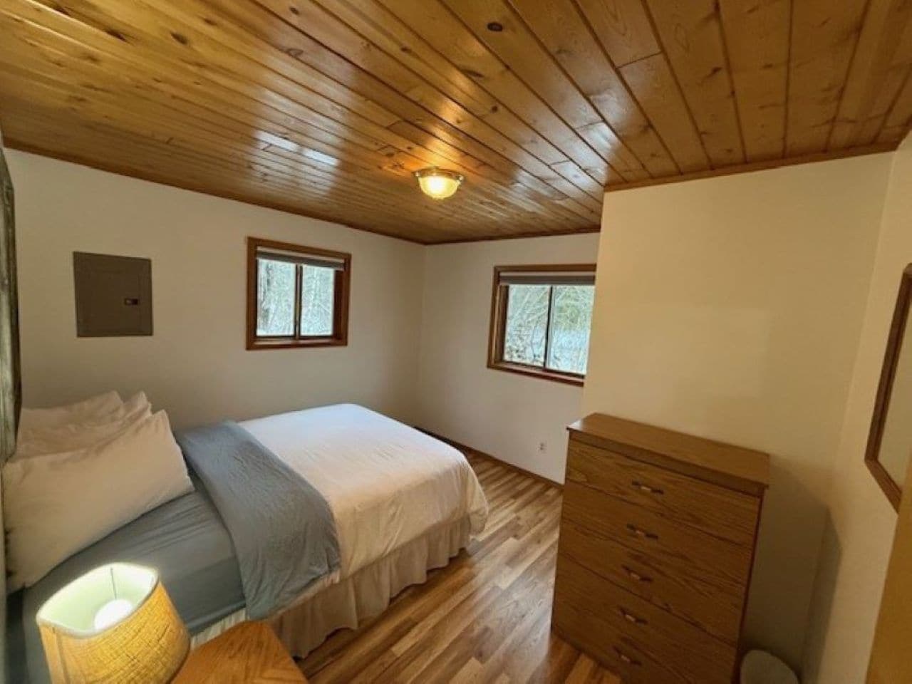 Une chambre avec un plafond en bois, un lit recouvert d’une courtepointe blanche et grise, deux fenêtres et une commode en bois.