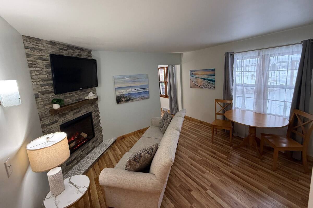 A living room with a stone fireplace, a flat-screen TV, a sofa, a dining table, and a window with curtains.