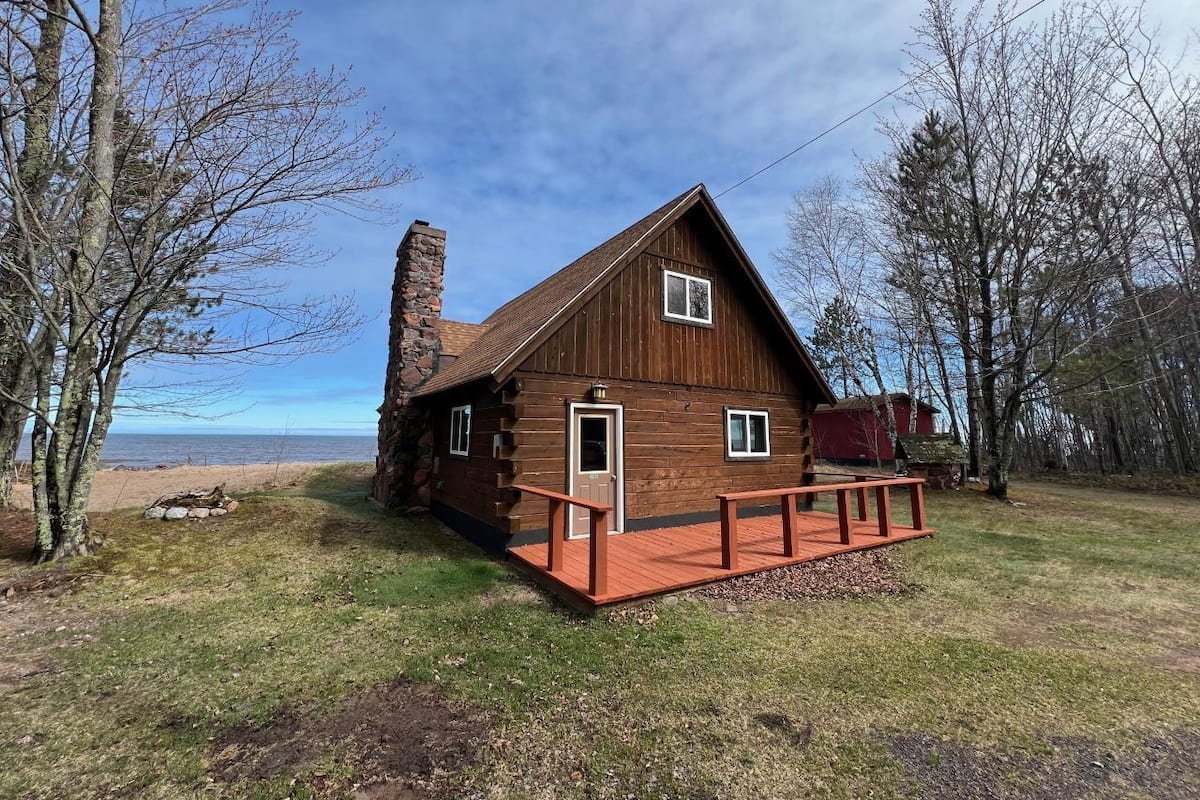 A wooden cabin with a stone chimney, a wooden deck, and a view of the beach.