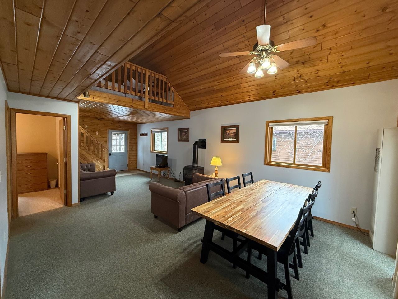 A spacious room with a wooden ceiling, a dining table with chairs, a sofa, a fireplace, and a staircase leading to an upper level.