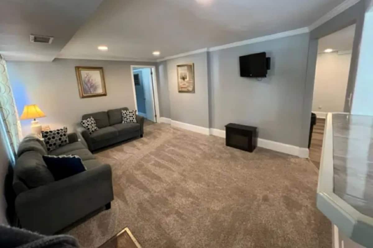 A living room with a grey sofa set, a flat-screen TV mounted on the wall, and a carpeted floor.